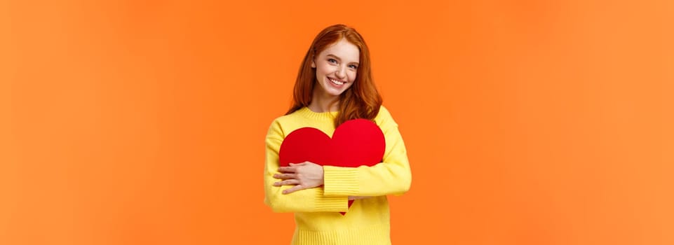 Valentines day, romance or relationship concept. Tender, lovely redhead girl waiting for her loved one, receive valentine card, hugging large heart sign and smiling happy, standing orange background.