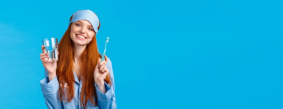 Happy smiling and carefree redhead enthusiastic girl in sleep mask, pyjama, holding glass water and toothbrush, taking care mouth hygiene, dental daily routine, blue background.