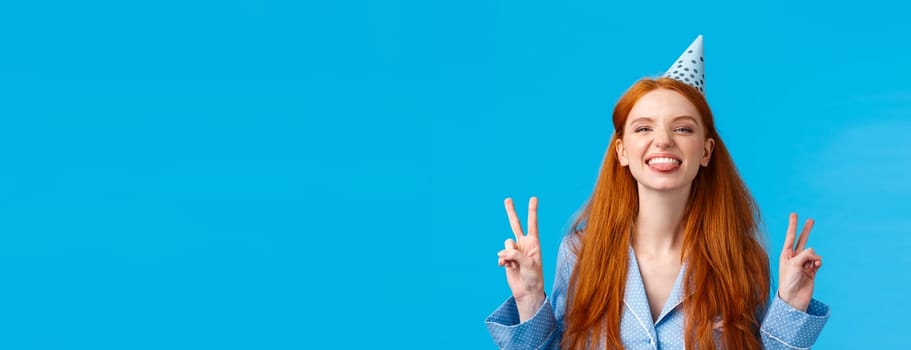 Finally eighteen. Cheerful and enthusiastic carefree redhead woman with long hair, freckles wearing nightwear, b-day cap, showing tongue happy, make peace gestures, standing blue background.