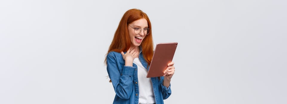Excited, happy and surprised cheerful redhead woman messaging with friend using gadget, holding digital tablet, touch chest pleased and amazed, laughing out loud looking device screen.