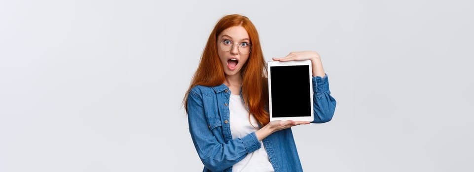 Excited and amused attractive redhead teenage girl making design, creative project on device screen, showing digital tablet display, look amazed, open mouth fascinated, white background.