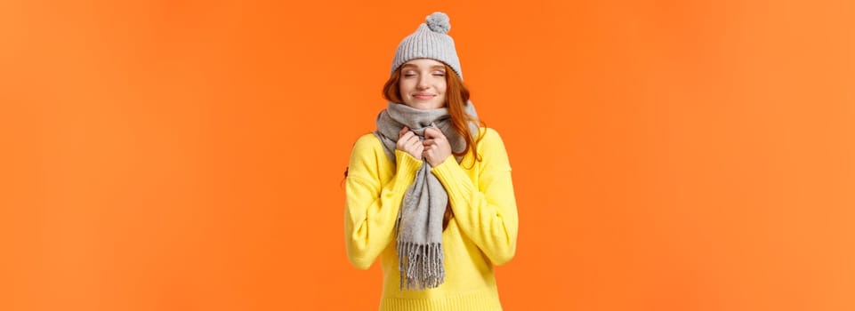 Cute and happy redhead woman walking along winter holidays fair market, close eyes and smiling as smelling something delicious, wearing warm grey hat and scarf, standing orange background.