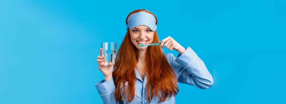 Enthusiastic upbeat, lively redhead woman, foxy girl with pretty smile, brushing teeth holding toothbrush and glass water, wake up morning, getting dressed, wearing sleep mask and pyjama.