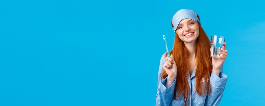 Happy smiling and carefree redhead enthusiastic girl in sleep mask, pyjama, holding glass water and toothbrush, taking care mouth hygiene, dental daily routine, blue background.