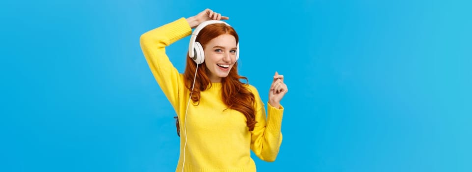 Cheeky and joyful, happy smiling redhead ecstatic woman having fun, listen music and lifting hands up as dancing, enjoying awesome sound quality, standing blue background delighted.