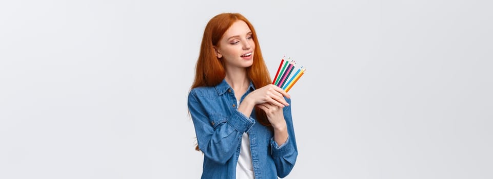 Creative and skilful good-looking redhead female in denim shirt, picking colored pencils, smiling thinking what draw, creating artworks, standing white background thoughtful.