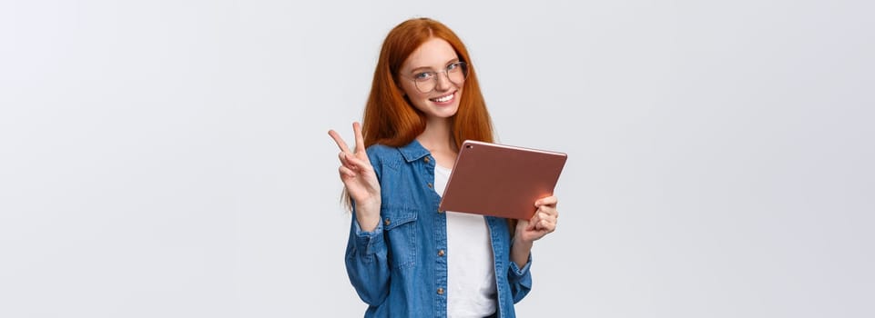 People, work and education concept. Cheerful attractive redhead female student in glasses, studying remote online courses, watching video on digital tablet, showing peace sign and smiling.