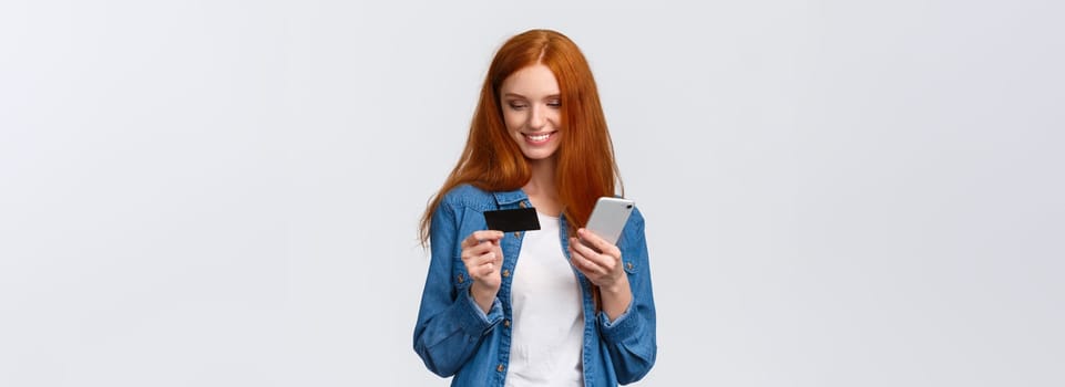 Finance, business and education concept. Cheerful cute alluring redhead woman making online purchase, holding smartphone and credit card, smiling as buying present for valentines day internet store.