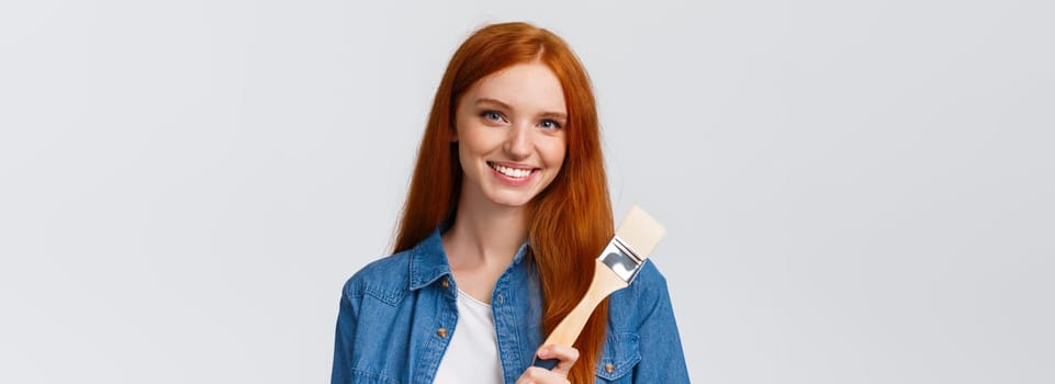 Close-up portrait talented and creative cute redhead girl with blue eyes like drawing, attend art courses, holding painting brush, consider paint walls new color, smiling confident, white background.