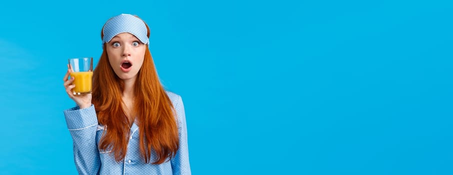 Amazed and speechless caucasian foxy woman in nightwear, pyjama and sleep mask, open mouth wondered, stare camera as holding orange juice, watching news morning breakfast, blue background.