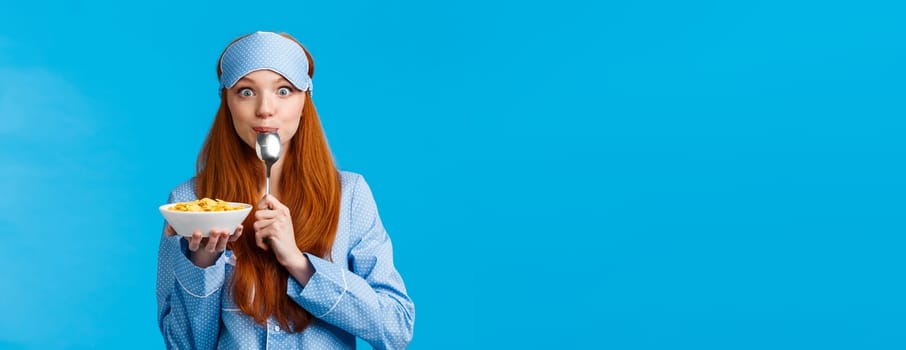 Mmm delicious. Cute pretty foxy teenager college girl licking spoon eating tasty cereals, holding plate, having breakfast wearing sleep mask and nightwear in morning, standing blue background.