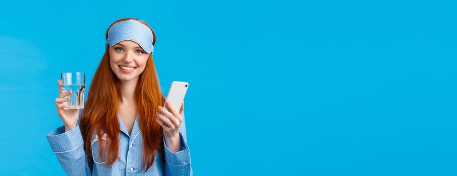 Waist-up portrait cheerful attractive glamour woman with red, ginger hair in sleep mask and pyjama, holding glass water and smartphone, smiling camera, standing blue background.