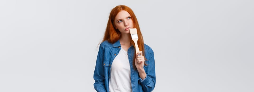 Thoughtful and creative redhead female imaging what colour paint room, holding paintbrush, look up thoughtful, thinking choices, pondering ideas, standing white background unsure.