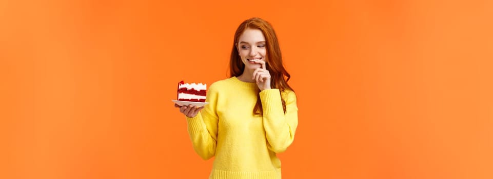 Cheerful and satisfied, happy redhead woman have cheat-day eating delicious food, holding tasty piece cake, biting lip and smiling, cant resist temptation, desire take bite, orange background.