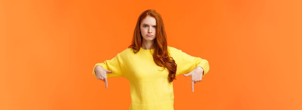 Mad unsatisfied cute redhead girlfriend, ginger girl in yellow sweater, frowning disappointed and angry, pointing down sulking, feeling offended with disapproval, standing orange background.