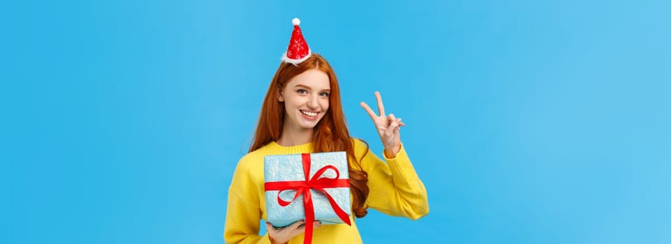 Waist-up portrait of cute happy redhead woman receive gift in wrapped box, celebrating christmas holidays, wearing fancy new year hat and showing peace sign, smiling joyfully, blue background.