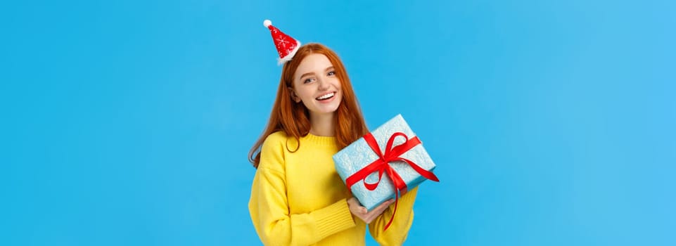 Holidays, celebration and presents concept. Cheerful redhead girl love christmas, enjoy celebrating with friends, receive gift in wrapped box from secret santa, wearing cute hat, blue background.