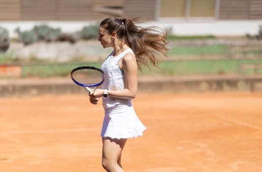 A woman in a white dress is holding a tennis racket and standing on a tennis court. She is focused and ready to play