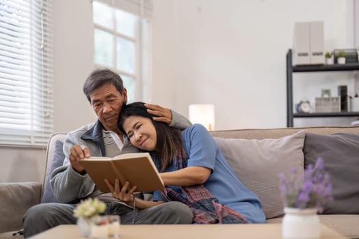 Elderly Asian couple enjoying a book together on a cozy couch, illustrating companionship and love in a home setting