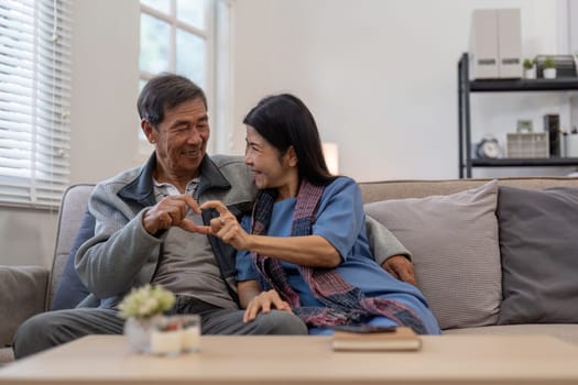 Healthy elderly Asian couple forming heart shape with their hands while sitting on a cozy couch, illustrating love and companionship in a home setting