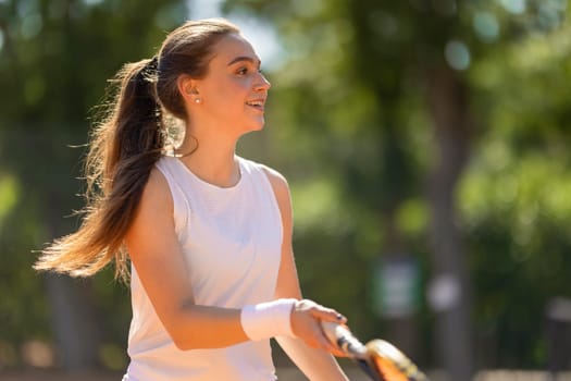 A woman is playing tennis and smiling. She is wearing a white tank top and a white wristband