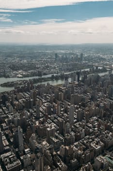 High-angle view of New York City showcasing numerous skyscrapers, the river, and vast cityscape under a partly cloudy sky. Image highlights the urban density and architectural diversity of the city.