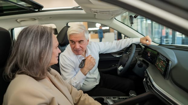 An elderly man holds the keys while sitting in a new car. A gray-haired married couple is happy about buying a car
