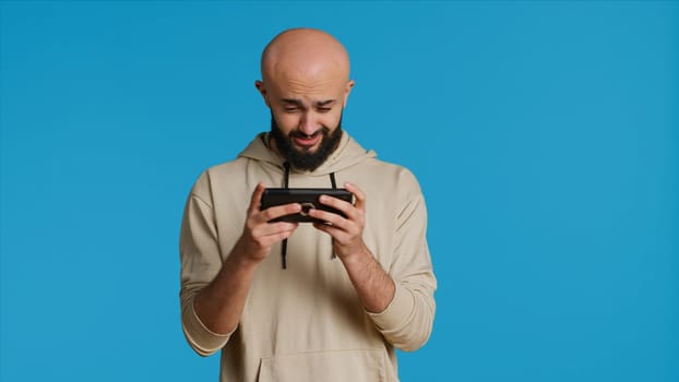 Joyful muslim man playing mobile video games in studio, enjoying online competition using smartphone app. Arabic gamer having fun with roleplaying challenge contest. Camera 1.