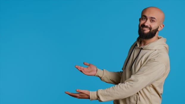 Smiling person advertising something on camera, pointing to left or right sides in studio. Middle eastern guy showing advertisement for marketing campaign, blue background pose. Camera 1.