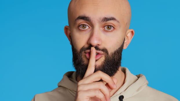 Middle eastern person showing mute gesture with finger over lips, posing over blue background. Arab guy doing hush symbol to keep silence, trying to share a secret whispering. Camera 2.