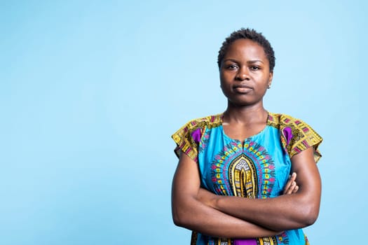 Portrait of young ethnic woman dressed in traditional clothing, posing with confidence in front of camera. African american girl in authentic colorful costume stands with arms crossed.