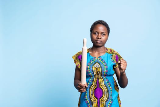 African american traditional woman holding rolling pin and threatening to fight someone in studio, wearing cultural attire. Female model acting serious and aggressive, grumpy girl.