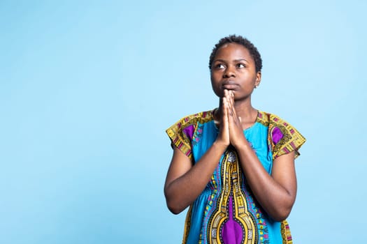 African american spiritual person holds hands in a prayer, praying to God in front of studio camera. Young woman with religious belief, worshipping Jesus over blue background.