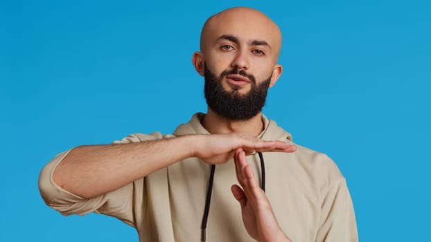 Middle eastern person doing timeout gesture on camera, asking for a break to stop working over blue background. Muslim adult presents symbol to pause or stop something. Camera 2. Handheld shot.
