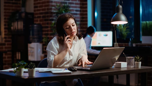 Woman answers phone call from husband while working in office at computer desk. Employee using smartphone to chat with friend during telephone call while at work in front of laptop at night, camera B