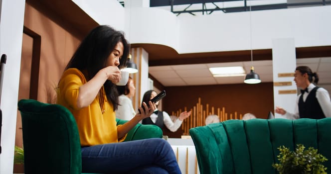 Tourist scrolling social media apps on smartphone, using free wifi connection in lounge area to pass time before doing check in. Asian woman relaxing on couch and planning trip itinerary.