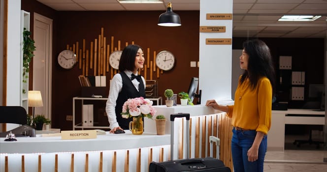 Receptionist welcoming asian woman at five star resort, asking about booking details to fill in registration forms. Front desk staff providing great services for client, presenting hotel amenities.