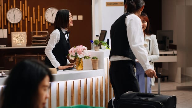 Asian traveler receiving passport and room access card to see accommodation after registration. Woman with luggage arriving at five star hotel, talking to receptionist at front desk.