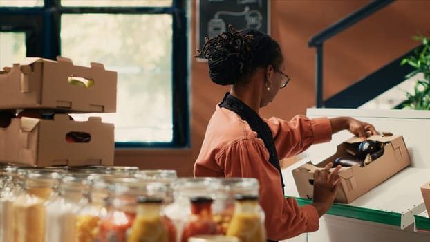 Merchant organizing fruits and veggies in crates on shelves, preparing fresh merchandise at farmers market before opening store. African american woman arranging natural produce.