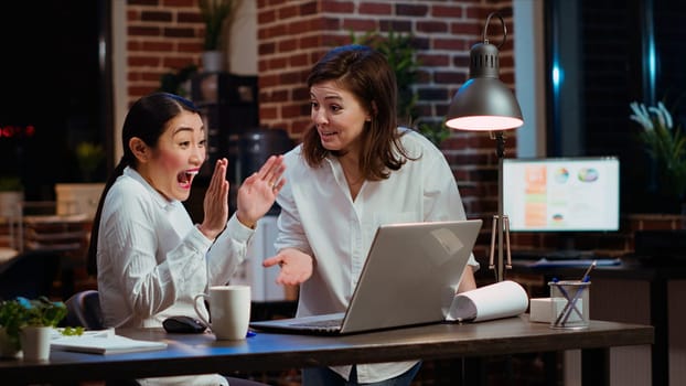 Jubilant businesswoman and coworker celebrating finalizing deal with customers after working together on laptop. Excited sales consultants doing high five after getting quality leads