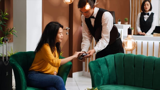 Asian client paying for coffee served in lounge area, using pos terminal payment to buy drink from hotel bar. Traveler making transaction with credit card upon her arrival at holiday retreat.