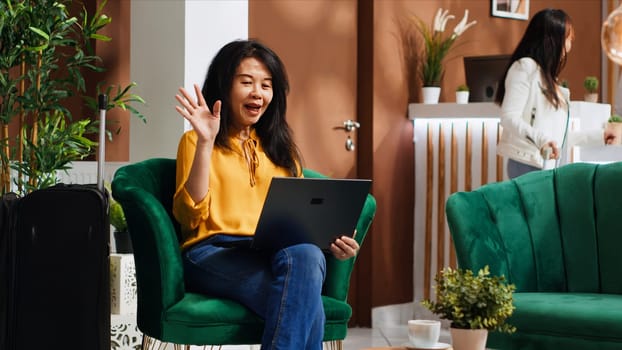 Hotel client waving on videocall connection with friends, talking to people after long international flight. Woman using laptop network to attend online teleconference remotely on trip. Handheld shot.