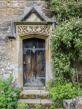 Entrance to a historic manor, framed by antique architectural elements and flanked by potted topiaries, features an aged door, the surrounding ivy and stonework add to the timeless elegance of the property