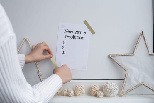 Unrecognizable woman creating vision board. NEW YEAR'S RESOLUTION checklist text on paper note stick on while wall with cozy minimalistic handmade Christmas decor. New year aims resolutions. Low key festive Planning and setting goals concept