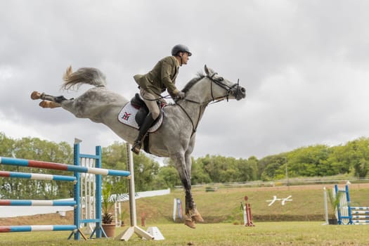 19 may 2024, Mafra, Portugal - competition in military academy - man is riding a horse and jumping over a hurdle. The horse is white and the man is wearing a green jacket. Concept of excitement and athleticism, as the man and horse are performing a challenging