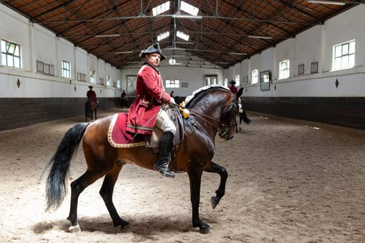 19 may 2024, Mafra, Portugal - competition in military academy - man in a red coat is riding a brown horse in a large, empty building. Scene is one of solitude and focus, as the man and horse are the only ones in the room