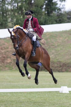 19 may 2024, Mafra, Portugal - competition in military academy - man in a red coat riding a horse. The horse is jumping over a white line. The man is wearing a hat and a vest