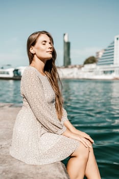 A woman with long hair is sitting on a dock by the water. She is wearing a white dress and she is looking at the water. The scene has a calm and peaceful mood