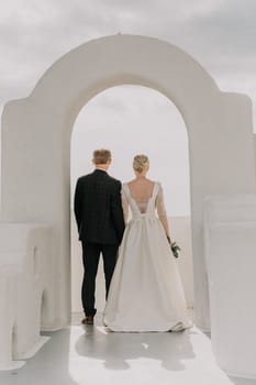 A bride and groom are walking through a white archway on a sunny day. The bride is wearing a white dress and the groom is wearing a suit. The archway is decorated with flowers