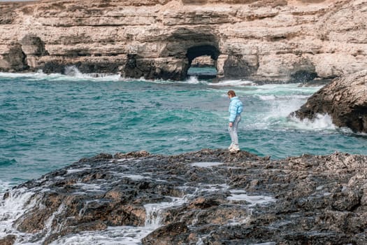 A woman in a blue jacket stands on a rock above a cliff above the sea and looks at the raging ocean. Girl traveler rests, thinks, dreams, enjoys nature. Peace and calm landscape, windy weather